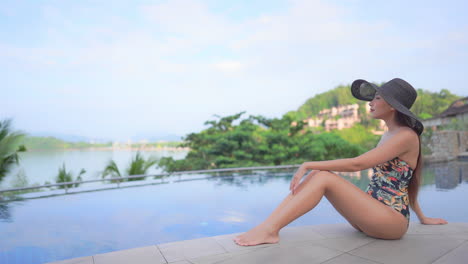 Lonely-Asian-Woman-in-Swimsuit-and-Floppy-Hat-Sitting-by-Swimming-Pool-Enjoying-in-Tropical-Landscape-View,-Full-Frame-Slow-Motion