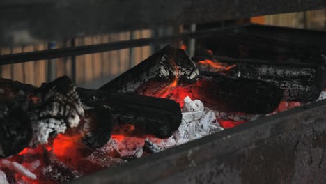 Slow-motion-pan-along-metal-barbecue-with-open-orange-hot-fire-flame-flickering-as-ash-and-embers-rise-up-from-smoke-with-red-and-white-charcoal-bed-in-rustic-outdoor-setting