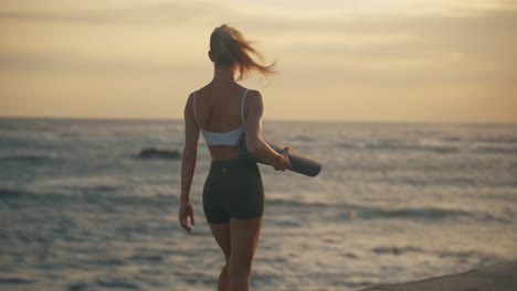 Fit-attractive-blond-woman-holds-yoga-mat-walking-along-shore,-golden-hour