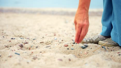 Elderly-Woman-Walking-On-The-Shore-And-Picks-Up-A-Stone-From-The-Sand-In-Karkle-Beach,-Lithuania---medium-shot