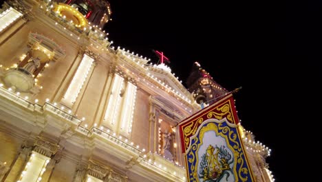 Malta,-Sliema,-religious-flag-hanging-in-front-of-a-decorated-church-during-a-local-festa-at-night