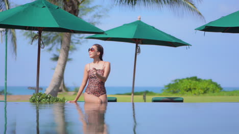 Lonely-sexy-exotic-woman-in-swimsuit-sitting-on-pool-border-with-tropical-landscape-in-background,-full-frame,-slow-motion