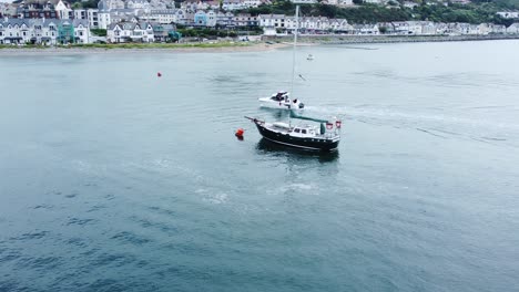 Power-boat-aerial-dolly-left-view-navigating-Conwy-marina-around-sailboat-with-mast-at-seaside-town