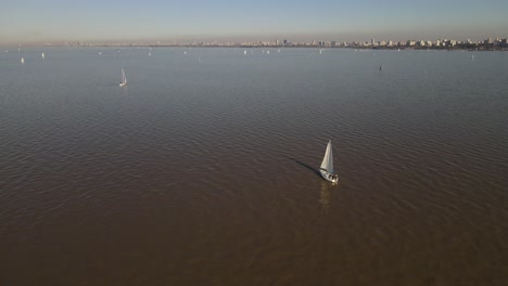 Beautiful-orbital-aerial-shot-with-a-small-white-sailing-boat-in-the-foreground-navigating-a-mighty-river-moving-towards-a-group-of-boats-with-a-modern-city-in-the-background