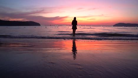 Eine-Silhouette-Eines-Mädchens,-Einer-Jungen-Frau-In-Einem-Kleid,-Die-Bei-Einem-Wunderschönen-Roten-Sonnenuntergang-Am-Strand-In-Richtung-Meer-Geht