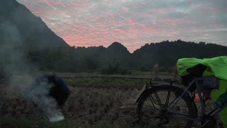 Junger-Asiatischer-Mann,-Der-Bei-Sonnenuntergang-Mit-Einem-Fahrrad-Ein-Lagerfeuer-Auf-Dem-Land-Macht