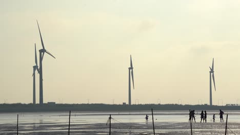 Silhouettes-of-tourists-walking-and-playing-on-tidal-flats-with-wind-turbines-spinning-at-the-background,-at-Gaomei-wetlands-preservation-area,-Taichung,-Taiwan