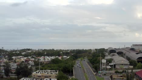 Aerial-descendent-view-over-Nunez-de-Caceres-street-in-Santo-Domingo-and-sea-in-background,-Dominican-Republic