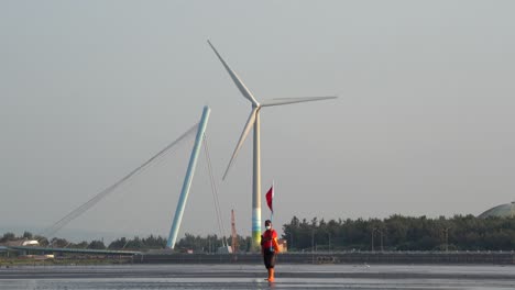 A-local-coastal-guard-holding-a-red-flag,-standing-still-with-a-giant-spinning-wind-turbine-and-bridge-in-the-background,-gaomei-wetland-preservation-area,-Taichung,-Taiwan