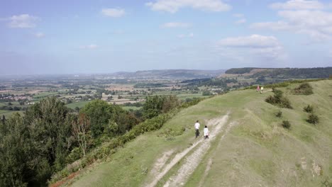 AERIAL---Couple-walk-their-dog-on-a-hill-near-Uley,-Cotswolds,-England,-forward