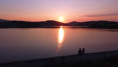Couple-sitting-on-the-wall-by-the-lake-during-sunset