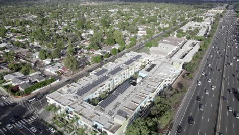 Pull-back-aerial,-Encino-apartment-complex,-solar-panels-on-roof,-over-101-Hollywood-freeway