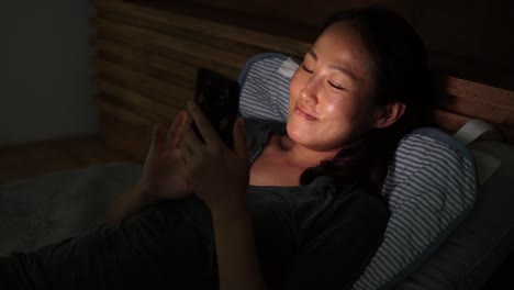 Mujer-Coreana-Asiática-Mirando-En-La-Pantalla-Del-Teléfono-Inteligente-Y-Sonriendo-Mientras-Descansa-En-La-Cama-Por-La-Noche-En-Plena-Oscuridad