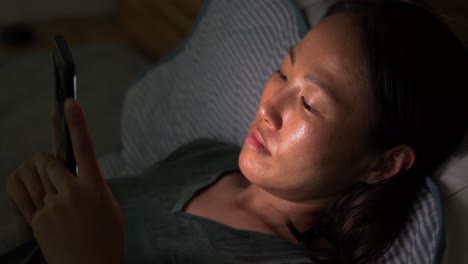 Koreanische-Frau-In-Ihrem-30.,-Die-Nachts-Auf-Dem-Telefonbildschirm-Mit-Ernstem-Gesicht-Zuschaut,-Während-Sie-Auf-Einem-Bett-In-Einem-Dunklen-Raum-Liegt