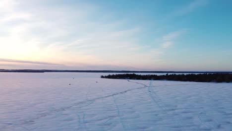 Frozen-snow-covered-lake-road-with-ice-fishing-huts,-ice-road-and-sled-during-sunset