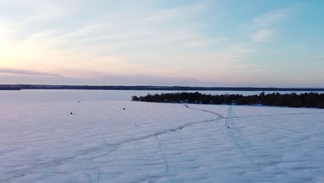 Frozen-snow-covered-lake-road-with-ice-fishing-huts,-ice-road-and-sled-during-sunset