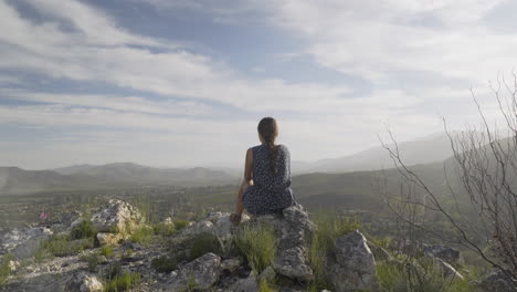 Girl-Sits-High-Up-On-A-Mountain-Looking-Down-On-A-Rural-Countryside-Town