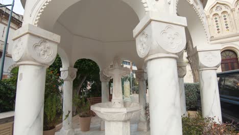 White-Marble-stone-Fountain-Gazebo-with-large-Cross-in-Agia-Napa-Cathedral,-Limassol,-Cyprus---Wide-Orbit-Tracking-shot