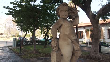 Statue-of-Eros-cupid-angel-ornament-embellishing-a-garden-in-Limassol,-Cyprus---Wide-Orbit-Tracking-shot