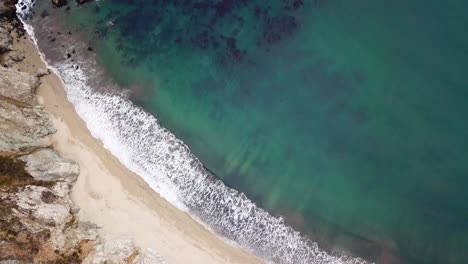 Turquoise-ocean-crashing-waves-onto-clean-sandy-beach-and-rocks,-top-down-aerial-view,-Bixby-Bridge-Seaside