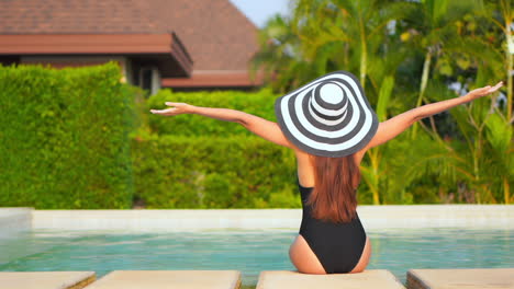 A-woman-with-her-back-to-the-camera-in-a-huge-sun-hat-and-swimsuit-sits-on-the-edge-of-a-swimming-pool-stretches-out-her-arms-in-welcome