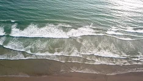 Aerial-birds-eye-of-waves-crashing-onto-a-dark-sandy-beach-with-sun-glaring-on-the-ocean