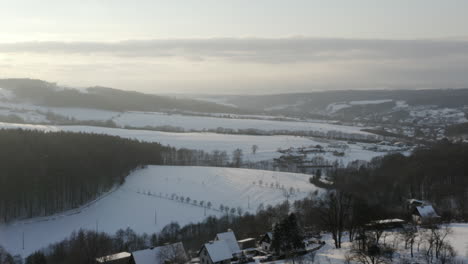 Sunrise-Aerial-Shot-Of-Farm-Village-During-Winter,-Snow-Covered-Field-And-Forest-Mountains