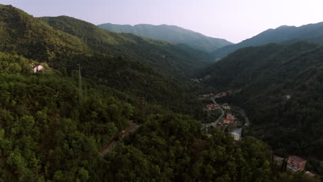 Drone-flying-towards-a-small-village-in-the-middle-of-a-valley-surrounded-by-forest,-black-car-driving-on-the-road-leading-towards-village-in-Italy