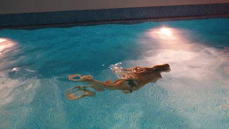 Slow-motion-shot-of-a-young-woman-swimming-inside-of-a-pool-during-night-time-by-herself,-dark-surroundings-and-pool-lights