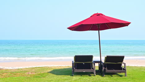 Roter-Sonnenschirm-Und-Liegestuhl-Auf-Grünem-Rasen-Nahe-Strandweiß-Mit-Meer-Im-Hintergrund