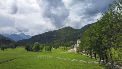Timelapse-of-storm-clouds-above-small-alpine-village-and-church-surrounded-by-meadows-and-tree-alley,-Zgornje-Jezersko,-Slovenia