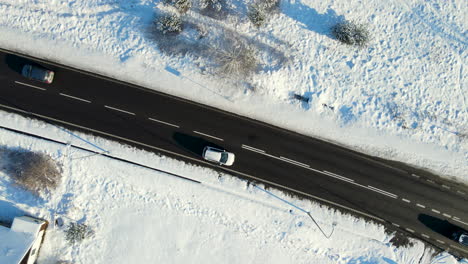 Aerial-orbit-shot-of-road-with-driving-cars-surrounded-by-winter-landscape-with-snow-and-snow-covered-trees-during-sunny-day