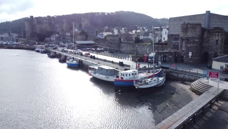 Idyllic-Conwy-castle-and-harbour-fishing-town-boats-on-coastal-waterfront-aerial-low-pull-back-over-pier