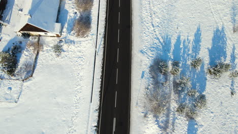 Aerial-top-to-down-view-drone-slowly-following-empty-winter-countryside-road-in-the-winter-landscape,-trees-fields,-and-houses-covered-with-snow