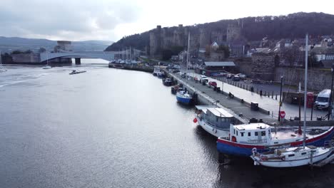 Idyllic-Welsh-Conwy-castle-and-harbour-fishing-town-boats-on-coastal-waterfront-aerial-fly-down-historic-landmark