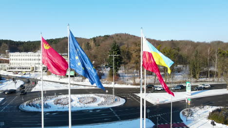 Slowly-flapping-European,-Poland-and-Ukraine-flags-with-at-the-background-cars-waiting-for-a-traffic-light-at-a-from-snow-cleared-roundabout