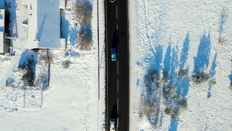 Aerial-top-down-of-cars-driving-on-black-asphalt-road-surrounded-by-white-winter-snowscape-during-sun