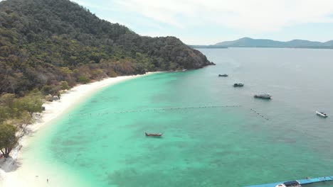 Lush-Green-steep-hill-and-sandy-shoreline-encircling-shallow-turquoise-sea-of-Koh-Hey-with-moored-boats-in-Thailand---Aerial-fly-over-rotating-orbit-shot