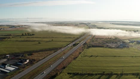 Patchy-dense-morning-fog-hangs-over-interstate-highway-with-traffic-passing-through-flat-green-farm-fields