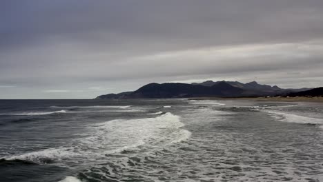 Aerial-backward-over-sea-waves-breaking-on-Rockaway-beach-with-cliffs-in-background