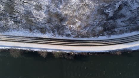 Birds-Eye-Aerial-View-of-Empty-Rural-Coutryside-Road-on-Winter-Season-Under-Snow-Capped-Cliffs-by-The-River