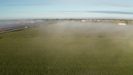 Vista-Aérea-De-La-Carretera-Con-Tráfico-Y-Cultivos-En-Campos-Al-Lado-De-La-Carretera-Con-Una-Fina-Capa-De-Niebla-Colgando-Del-Paisaje