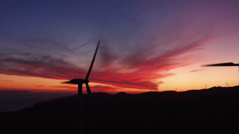 Aerial-close-up-silhouette-of-rotating-windmill-turbines,-producing-green-energy-outdoors-during-beautiful-sunset-in-the-evening