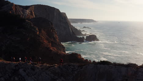 Aerial:-Silhouette-of-hiker-group-walking-on-narrow-trail-with-beautiful-coastline-with-crashing-atlantic-ocean-during-sunset