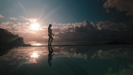 Woman-walking-beside-calm-infinity-pool-with-her-silhouetted-reflection-against-the-clouds-and-sun