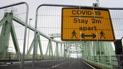 Covid-social-distancing-metal-sign-on-empty-closed-Runcorn-Jubilee-bridge-mesh-fence-dolly-left
