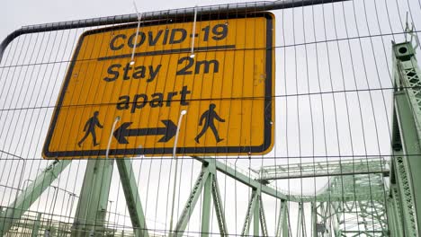 Covid-social-distancing-metal-sign-on-empty-closed-Runcorn-Jubilee-bridge-mesh-fence-close-dolly-right