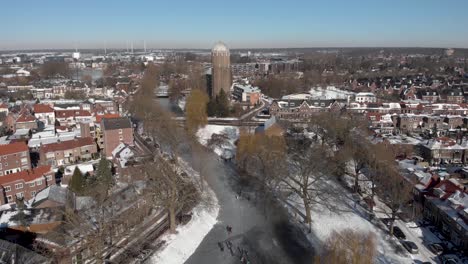 Ascending-aerial-winter-scene-with-people-ice-skating-along-the-curved-frozen-canal-going-through-the-Dutch-city-of-Zutphen-with-shadows-of-barren-trees-and-former-water-tower-in-the-background