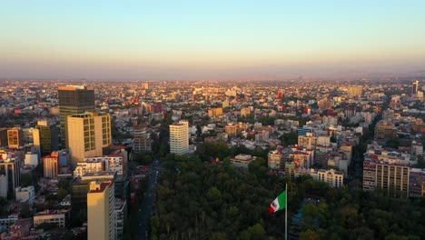 Puesta-De-Sol-Naranja-Vibrante-Sobre-La-Ciudad-De-México,-Parque-Hundido,-Bandera-Mexicana-Con-Retroceso-Aéreo-Ondeando