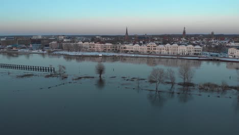 Aerial-cityscape-approach-of-Dutch-Hanseatic-medieval-tower-town-Zutphen-in-The-Netherlands-with-snow-on-the-boulevard-at-sunset-reflecting-with-vegetation-rising-above-overflown-river-IJssel